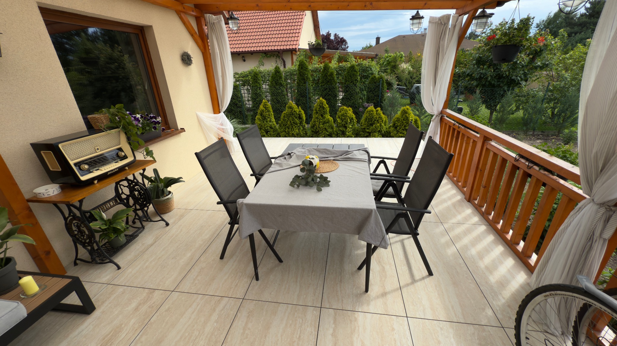 Zadaszony taras z beżową podłogą. Stół z krzesłami, dekoracje, radio na stoliku ze starej maszyny do szycia. W tle zielony ogród i białe zasłony.