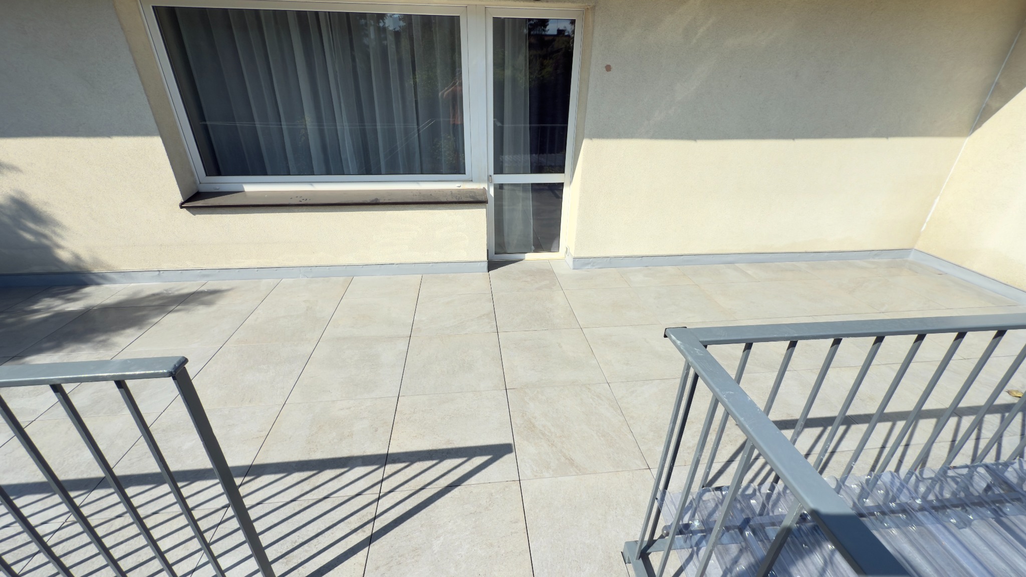 Jasny taras wyłożony kwadratowymi płytkami. Widoczna szara balustrada i okno z zasłoną. Świeże, minimalistyczne wykończenie.