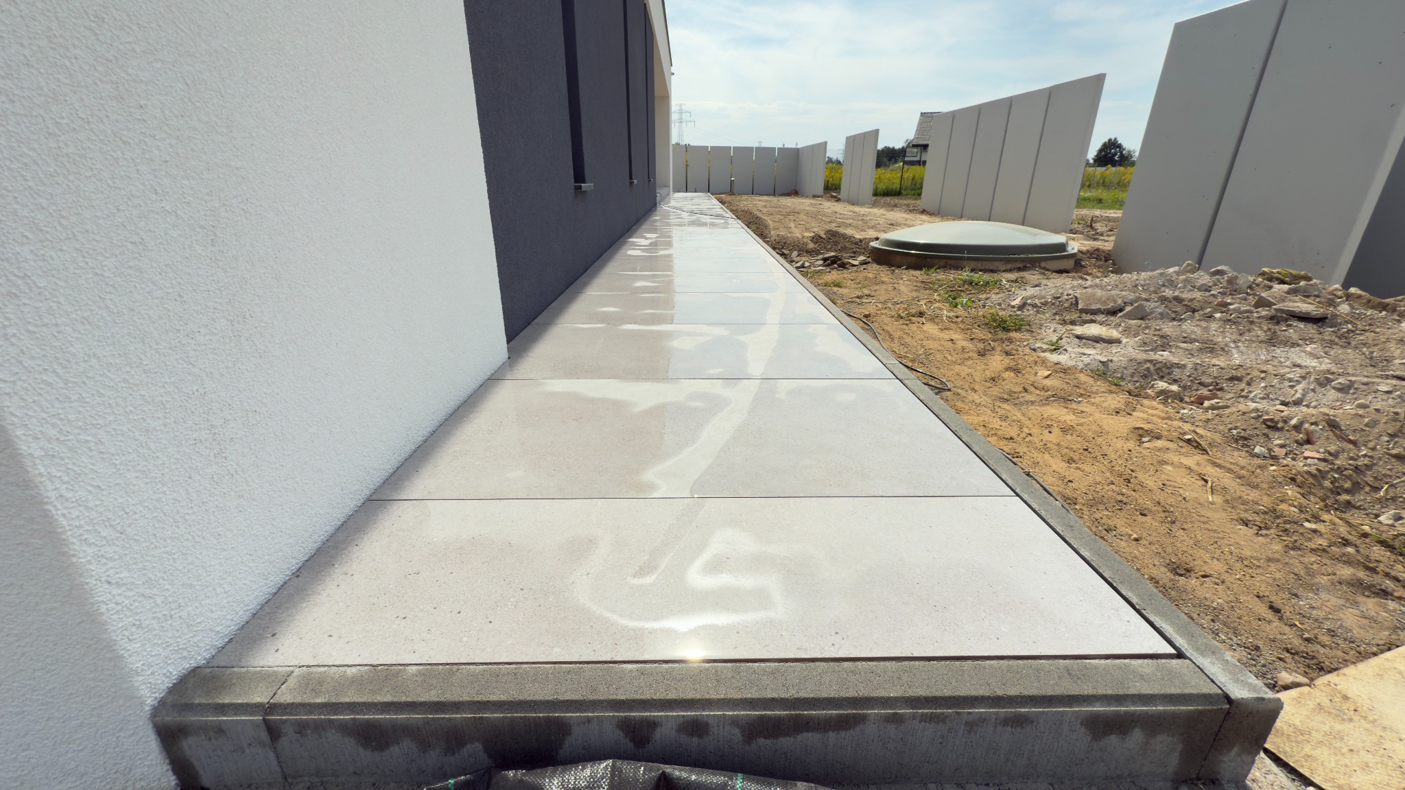 Nowoczesny taras z jasnych płyt betonowych przylegający do budynku, z widocznymi elementami wykończeniowymi i przygotowanym terenem wokół.