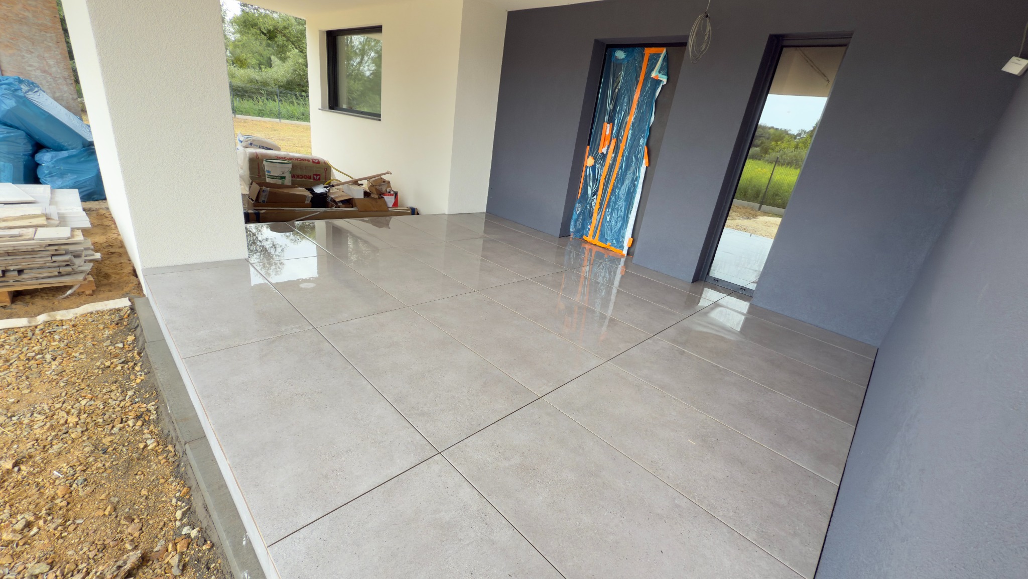 Jasne, kwadratowe płytki tarasowe, świeżo ułożone na zewnątrz budynku. Widoczne materiały budowlane i zabezpieczone folią drzwi. Dyskretne oświetlenie.