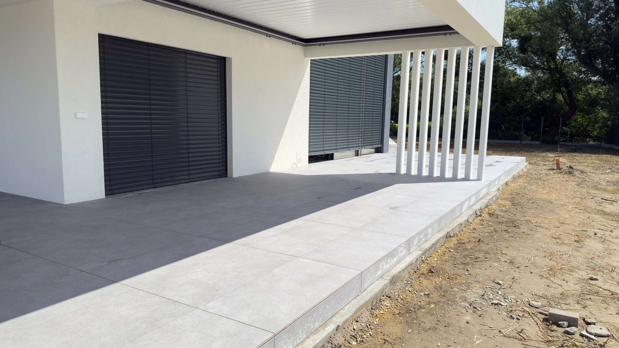 Nowoczesny taras z szarych płyt, zadaszony, z białymi, pionowymi elementami dekoracyjnymi. Widoczne rolety zewnętrzne i surowe otoczenie.