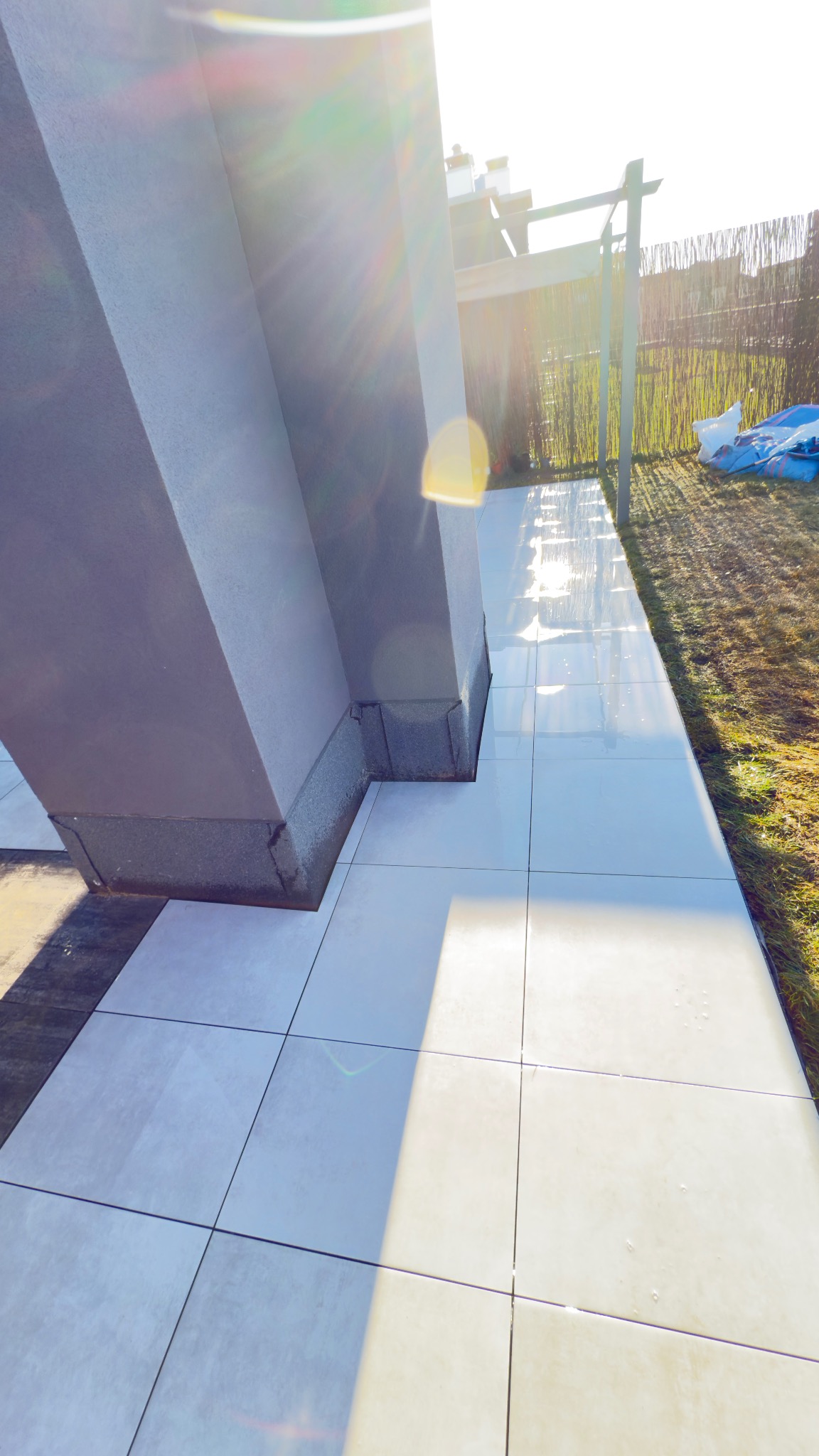 Jasne, kwadratowe płyty tarasowe ułożone przy szarej kolumnie, z widoczną zielenią w tle. Słoneczne światło odbija się od powierzchni płytek.