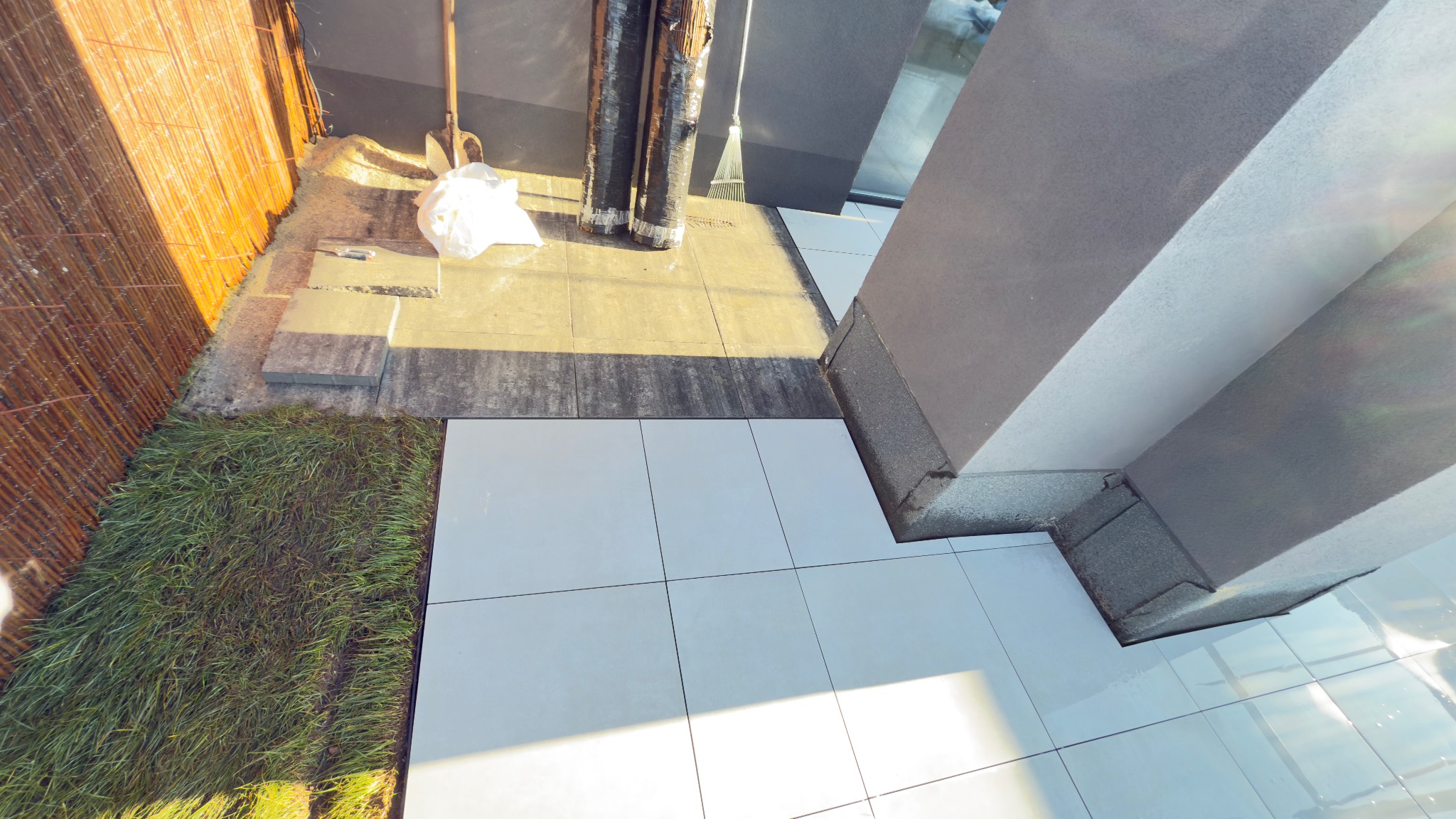 Układanie jasnych płytek na tarasie, widoczne materiały budowlane i fragment trawnika. Ujęcie z góry, w trakcie prac wykończeniowych.