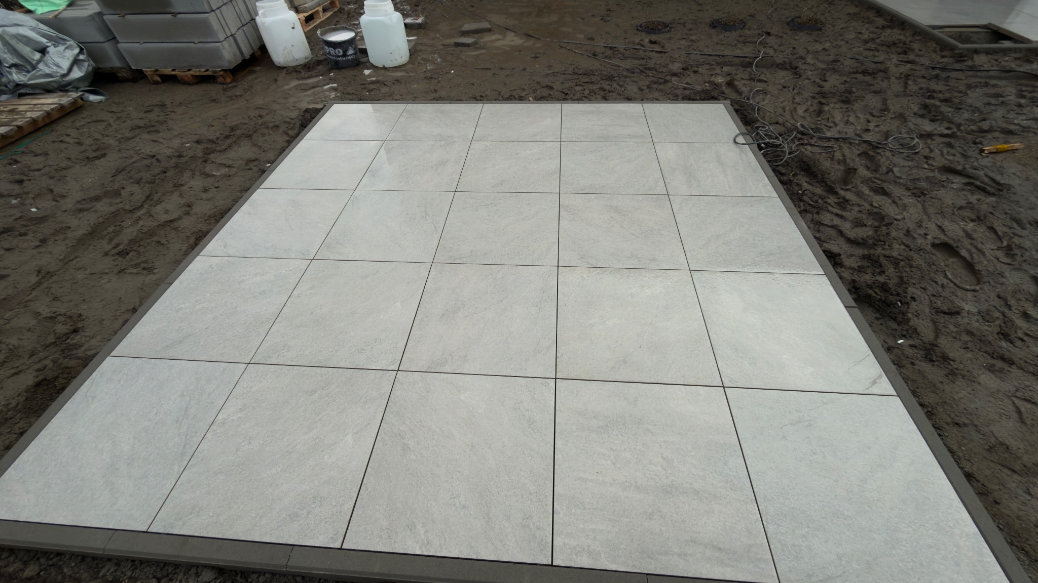 Jasnoszara, kwadratowa nawierzchnia z płyt ułożona na gruncie, obramowana ciemniejszymi elementami. W tle widoczne materiały budowlane i błoto.