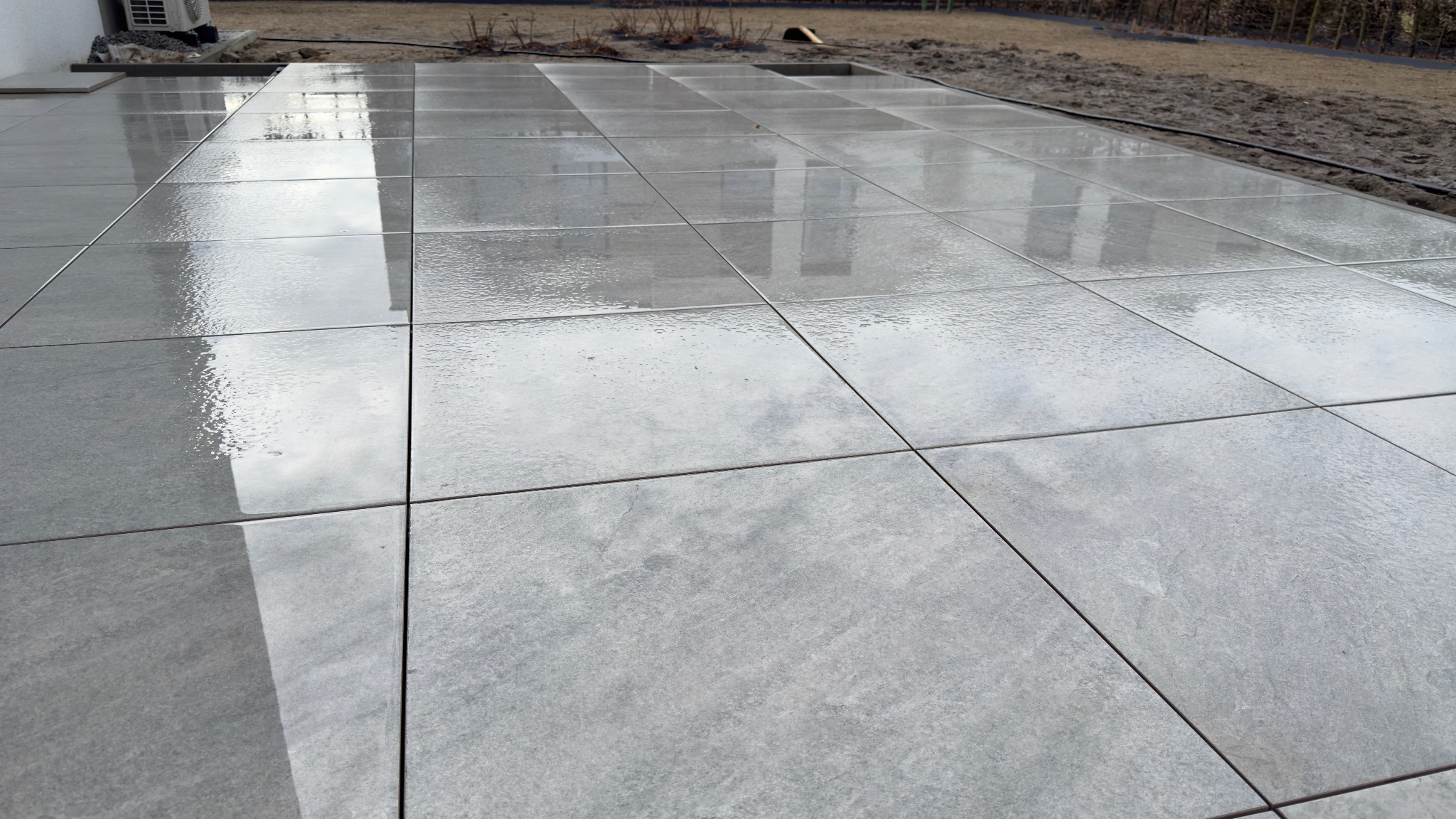 Świeżo ułożony, mokry taras z szarych, kwadratowych płyt betonowych. Widoczne odbicia nieba i otoczenia na powierzchni. Minimalistyczny design, równomierne fugi.