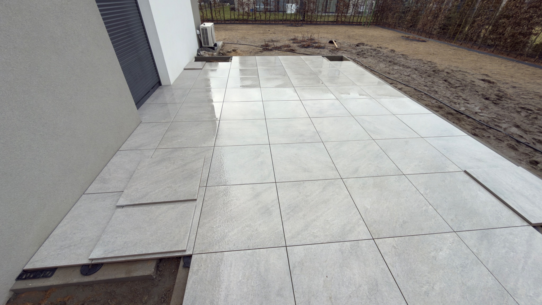 Jasnoszary taras z płytek w trakcie układania, widoczne elementy montażowe. W tle trawnik i krzewy, obok ściana budynku z klimatyzatorem.