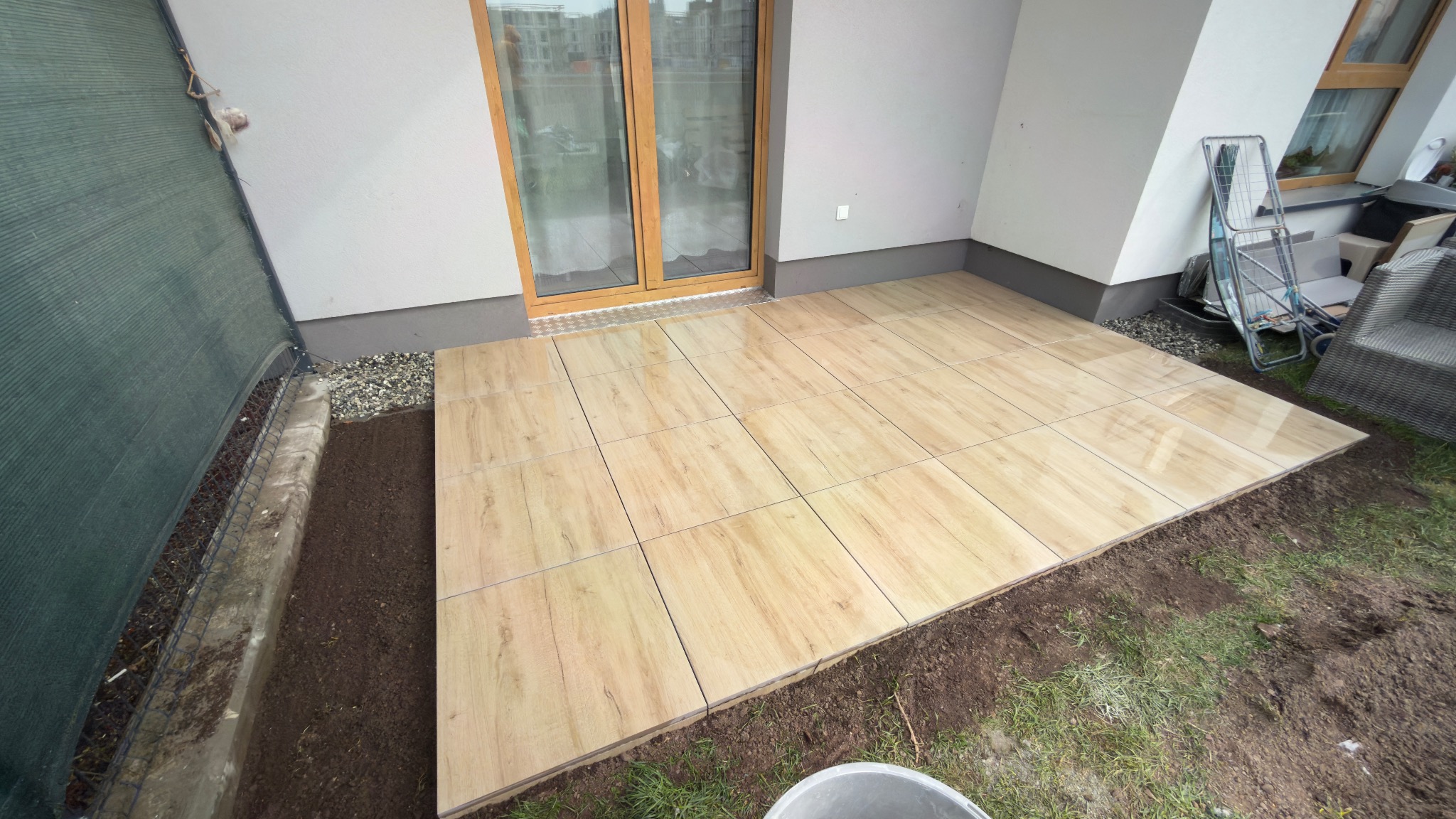 Ułożony taras z płytek imitujących drewno, przylegający do budynku z oknem balkonowym. Widoczne przygotowane podłoże pod trawnik i elementy wyposażenia.