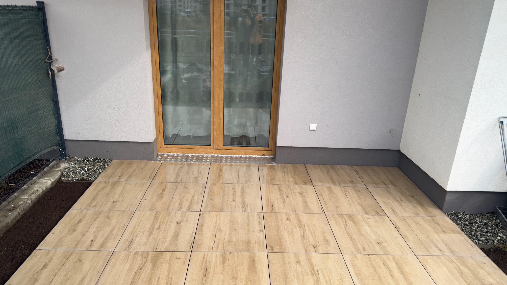 Jasne, drewnopodobne płytki tarasowe ułożone na balkonie, widok z góry. Fragment siatki ogrodzeniowej i okna balkonowe w tle. Minimalistyczny design.