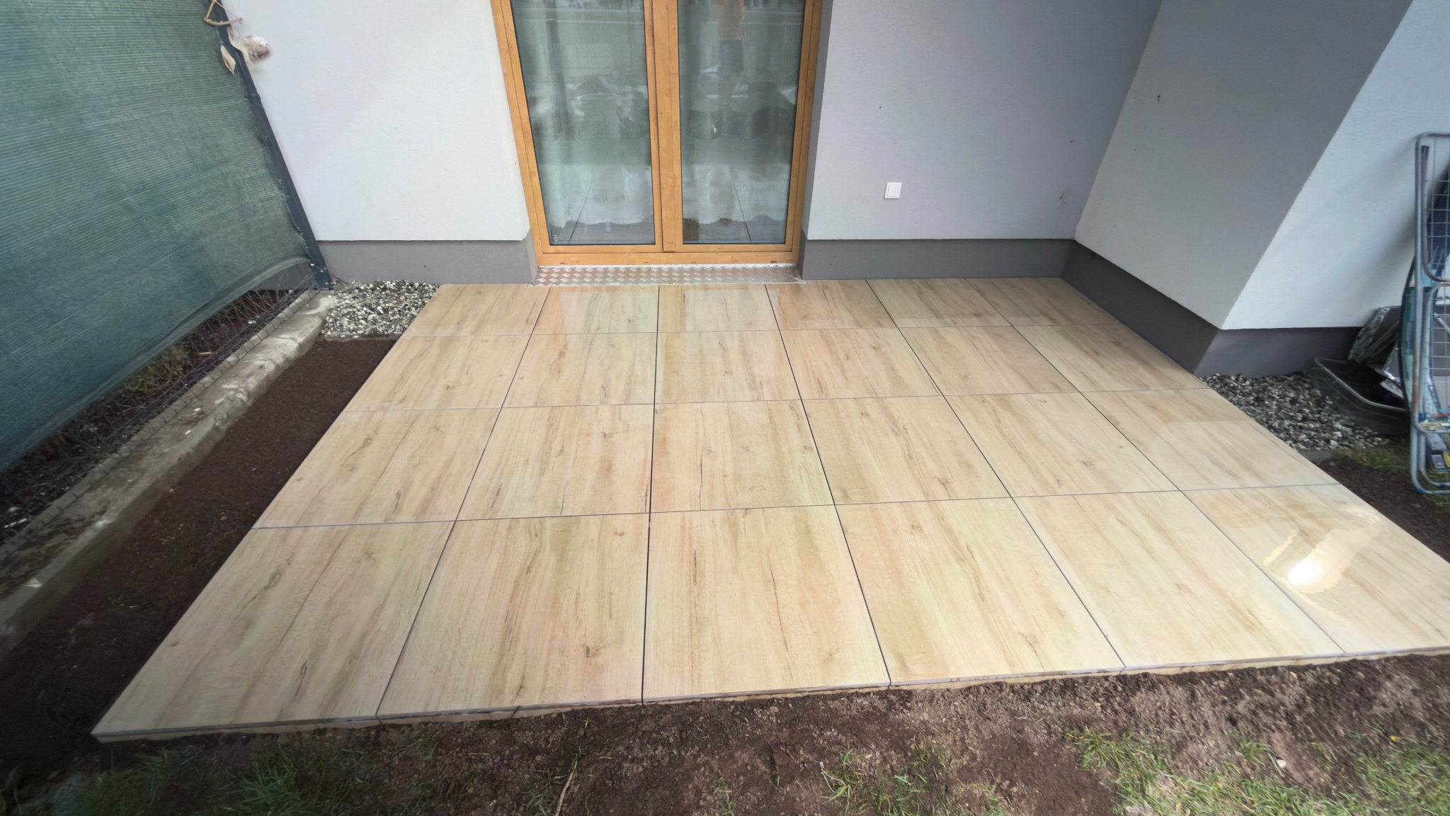 Jasne płytki imitujące drewno tworzą nowy taras przy domu. Widoczne świeżo obsypane ziemią obrzeża i siatka cieniująca. Ujęcie z niskiej perspektywy.