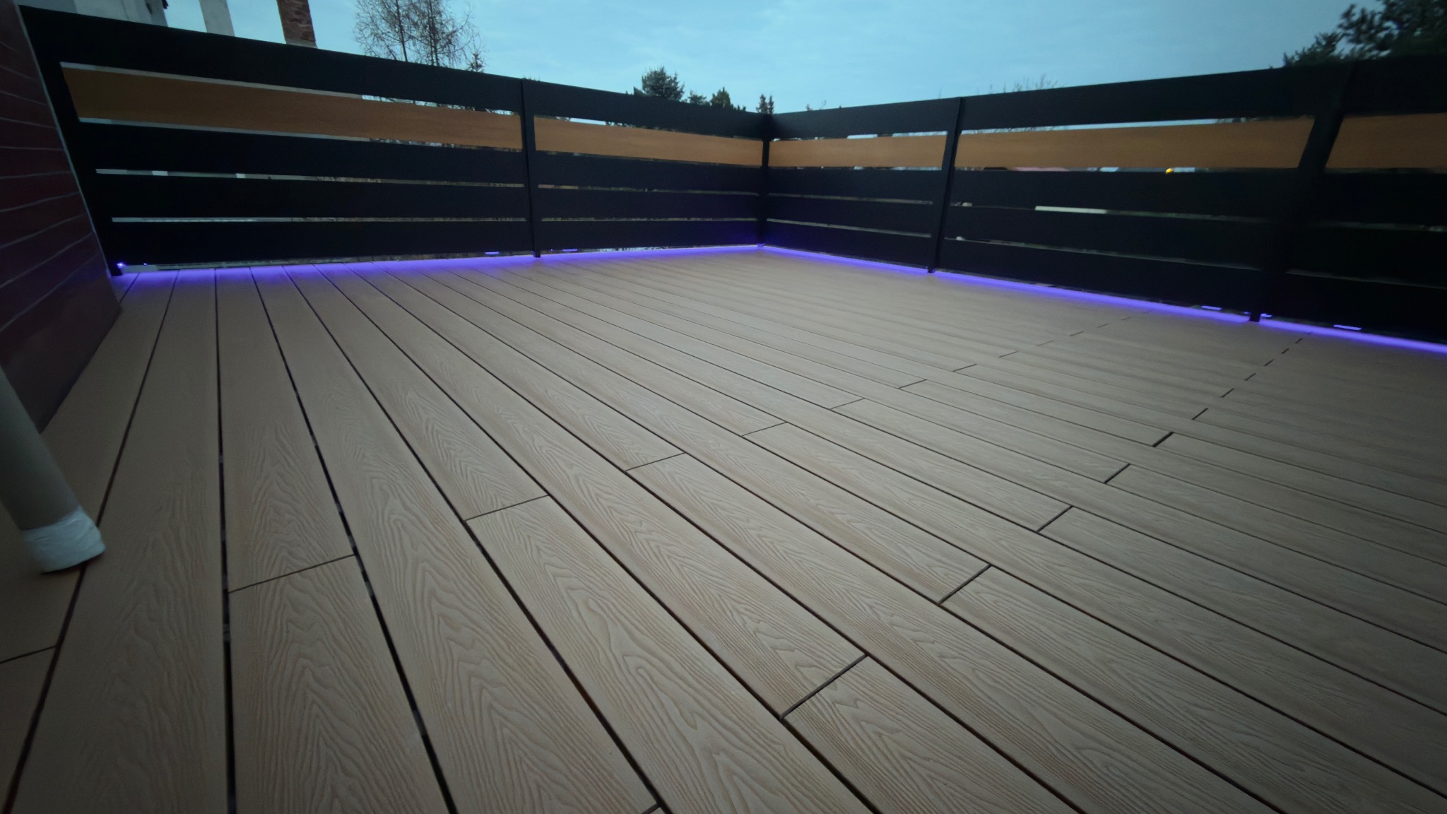 Nowoczesny taras kompozytowy z podświetleniem LED wzdłuż balustrady. Deski ułożone równolegle, widoczne łączenia. Balustrada czarna z drewnianymi elementami.