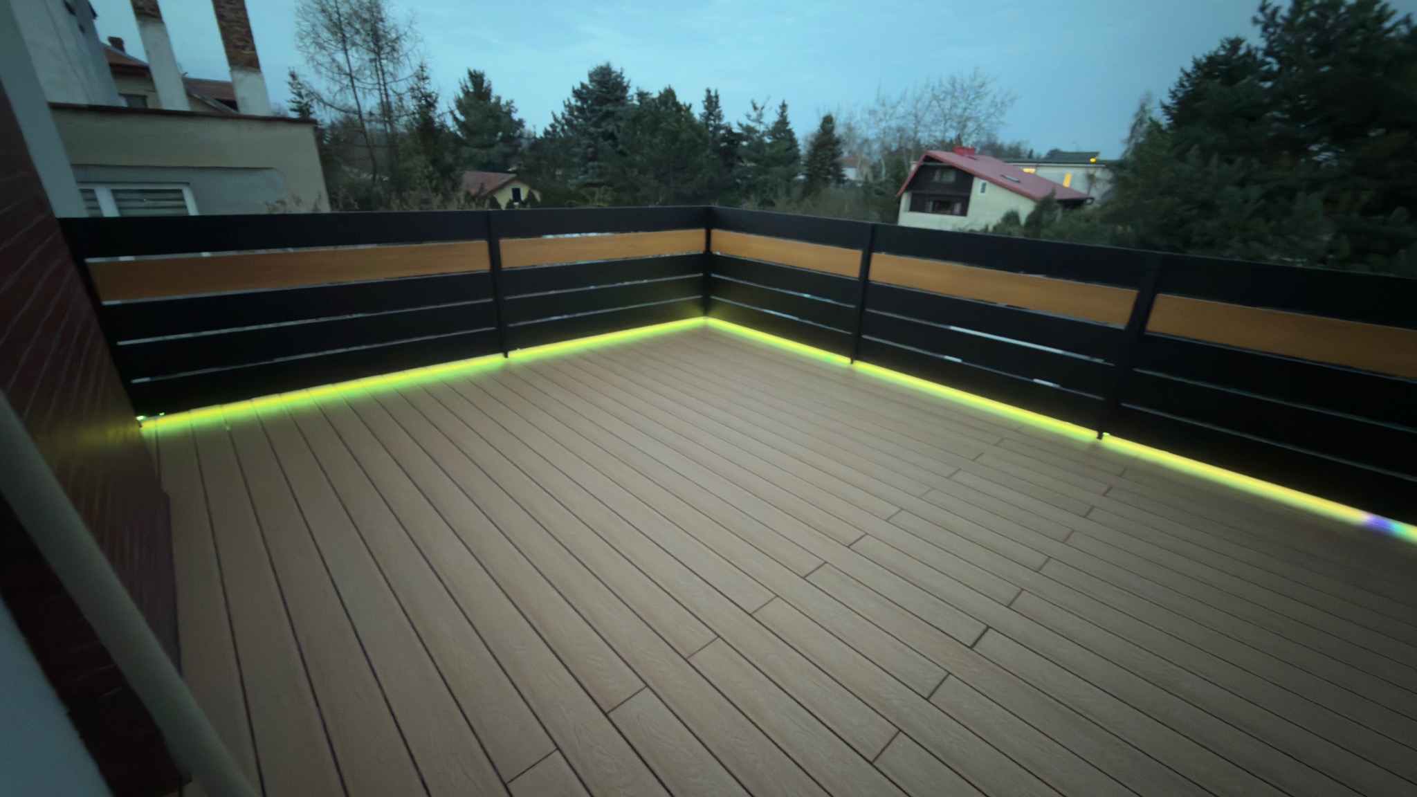 Nowoczesny taras kompozytowy z podświetleniem LED wzdłuż balustrady. Deski ułożone równolegle, widok na zieleń i zabudowania w tle. Wieczorny klimat.