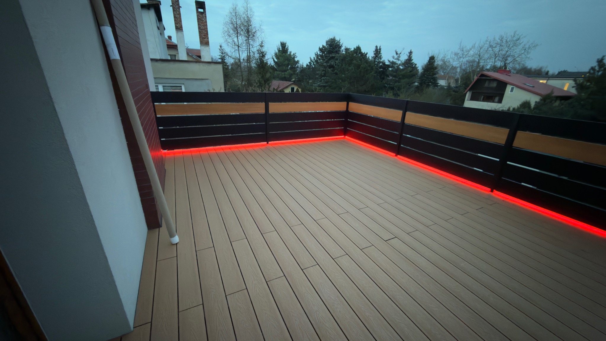 Nowoczesny taras kompozytowy z podświetleniem LED wzdłuż balustrady, widok z góry, w tle zabudowa jednorodzinna i zieleń. Ujęcie z niskiego kąta.