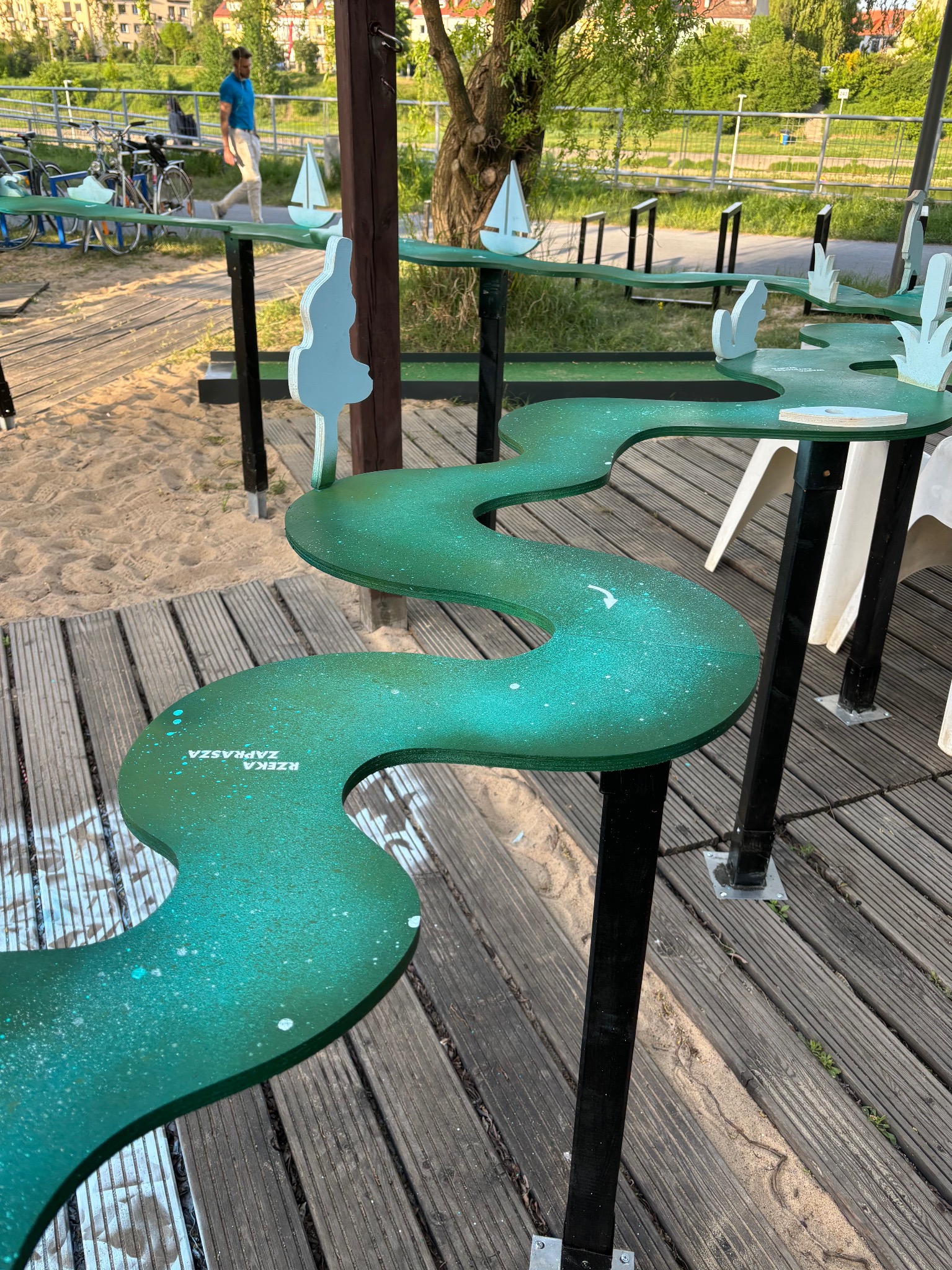 Zielony, kręty tor z elementami dekoracyjnymi w kształcie drzew i żaglówek, umieszczony na drewnianym podłożu, z napisem 'Rzeka Zaprasza'.