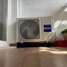 ENGETIA - Zewnętrzna jednostka klimatyzacji Haier, umieszczona na czarnych podkładkach antywibracyjnych na balkonie, z widocznym przewodem odprowadzającym skropliny do czerwonej miski.