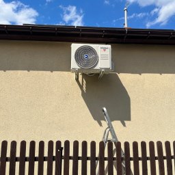 ENGETIA - Biała jednostka zewnętrzna klimatyzacji marki Kaisai zamontowana na elewacji budynku, widoczna drabina oparta o ścianę, brązowe ogrodzenie w dolnej części kadru, błękitne niebo z chmurami w tle.