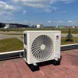 ENGETIA - Zewnętrzna jednostka klimatyzacji marki Gree, zamontowana na dachu przemysłowego budynku, widoczna z bliska, z tłem błękitnego nieba i zabudowań przemysłowych.