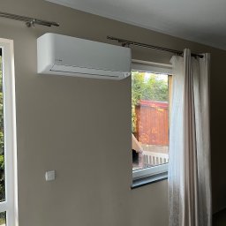 ENGETIA - Biały klimatyzator Rotenso zamontowany na ścianie w pokoju z beżowymi ścianami, widok na ogród przez okno z zasłonami.