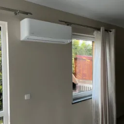 Biały klimatyzator Rotenso zamontowany na ścianie w pokoju z beżowymi ścianami, widok na ogród przez okno z zasłonami.