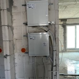 ENGETIA - Dwa białe panele elektryczne zamontowane na surowej ścianie z betonu komórkowego, widoczne okablowanie, surowy stan wykończenia wnętrza.