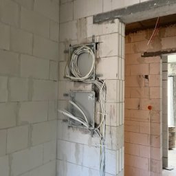 ENGETIA - Dwie puszki instalacyjne z kablami wychodzącymi ze ściany z betonu komórkowego, widok na sąsiednie pomieszczenie w stanie surowym.