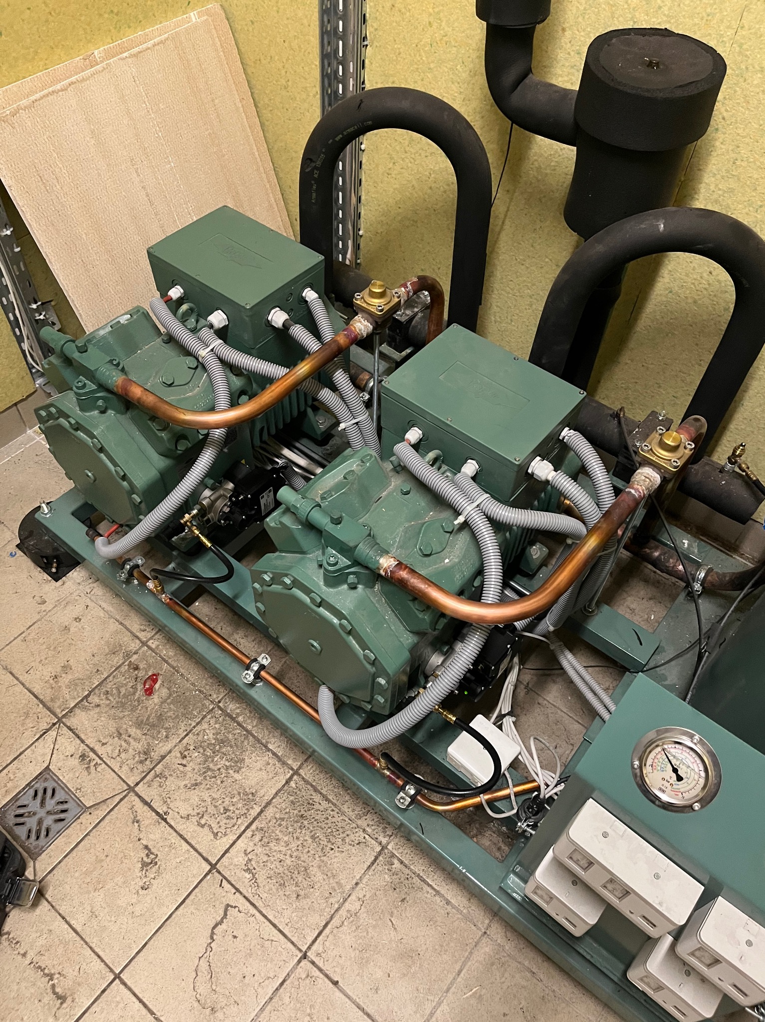 Kompresor chłodniczy w odcieniach zieleni z miedzianymi rurami i czarnymi izolowanymi przewodami, zainstalowany na podłodze z płytek ceramicznych, widoczny manometr.