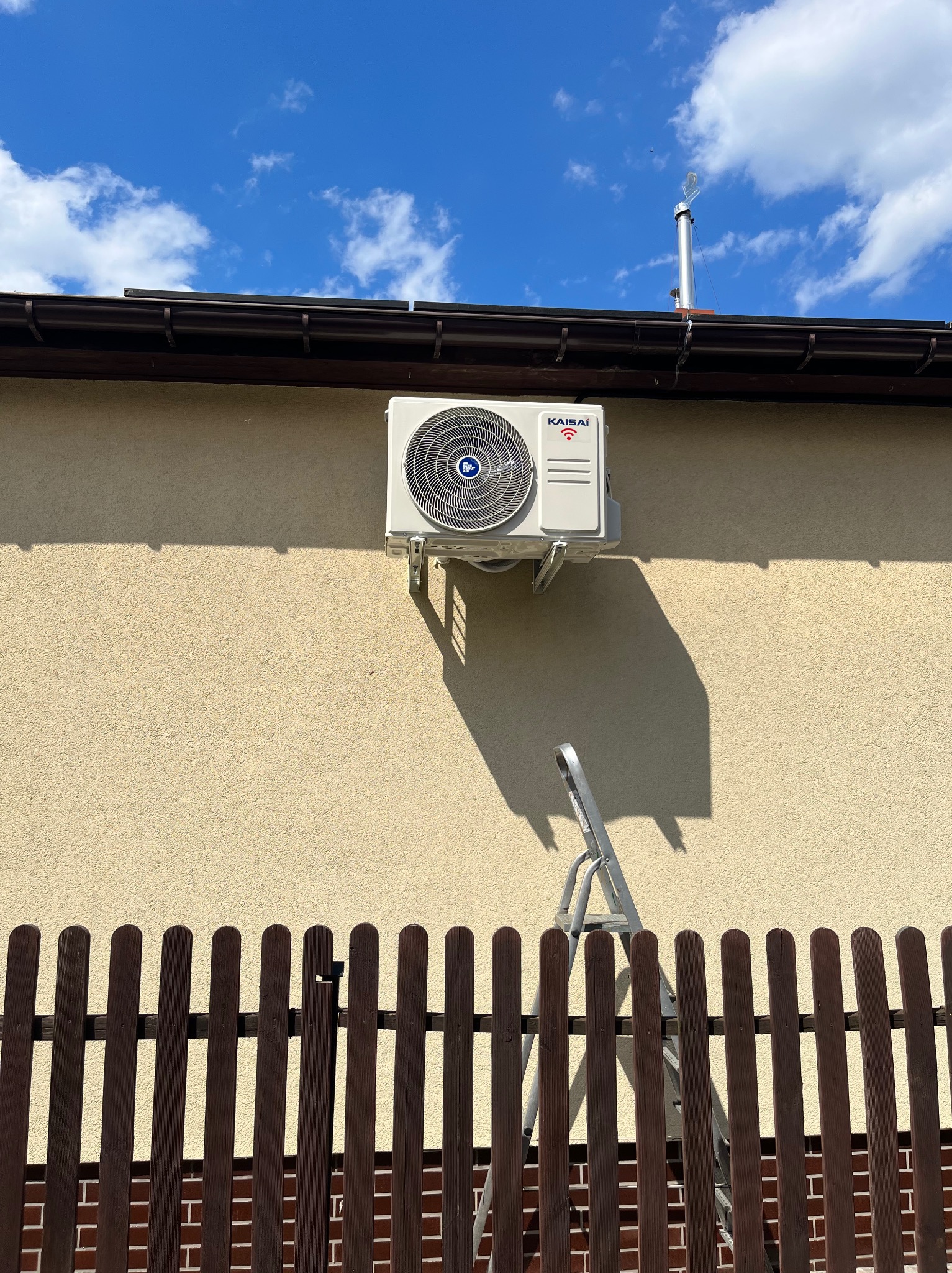 Biała jednostka zewnętrzna klimatyzacji marki Kaisai zamontowana na elewacji budynku, widoczna drabina oparta o ścianę, brązowe ogrodzenie w dolnej części kadru, błękitne niebo z chmurami w tle.