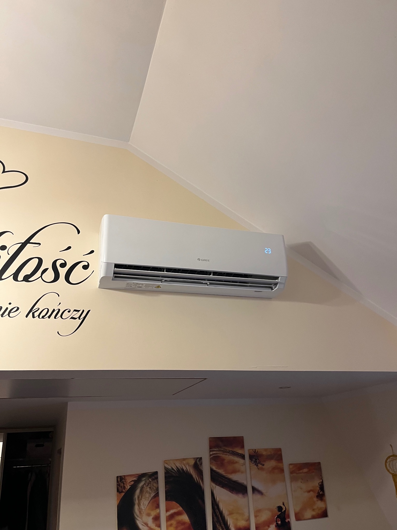 Biała klimatyzacja marki Gree zamontowana na ścianie w pomieszczeniu w Chojnie, nad nią skośny sufit, obok naklejka ścienna z napisem 'Miłość się nie kończy'.