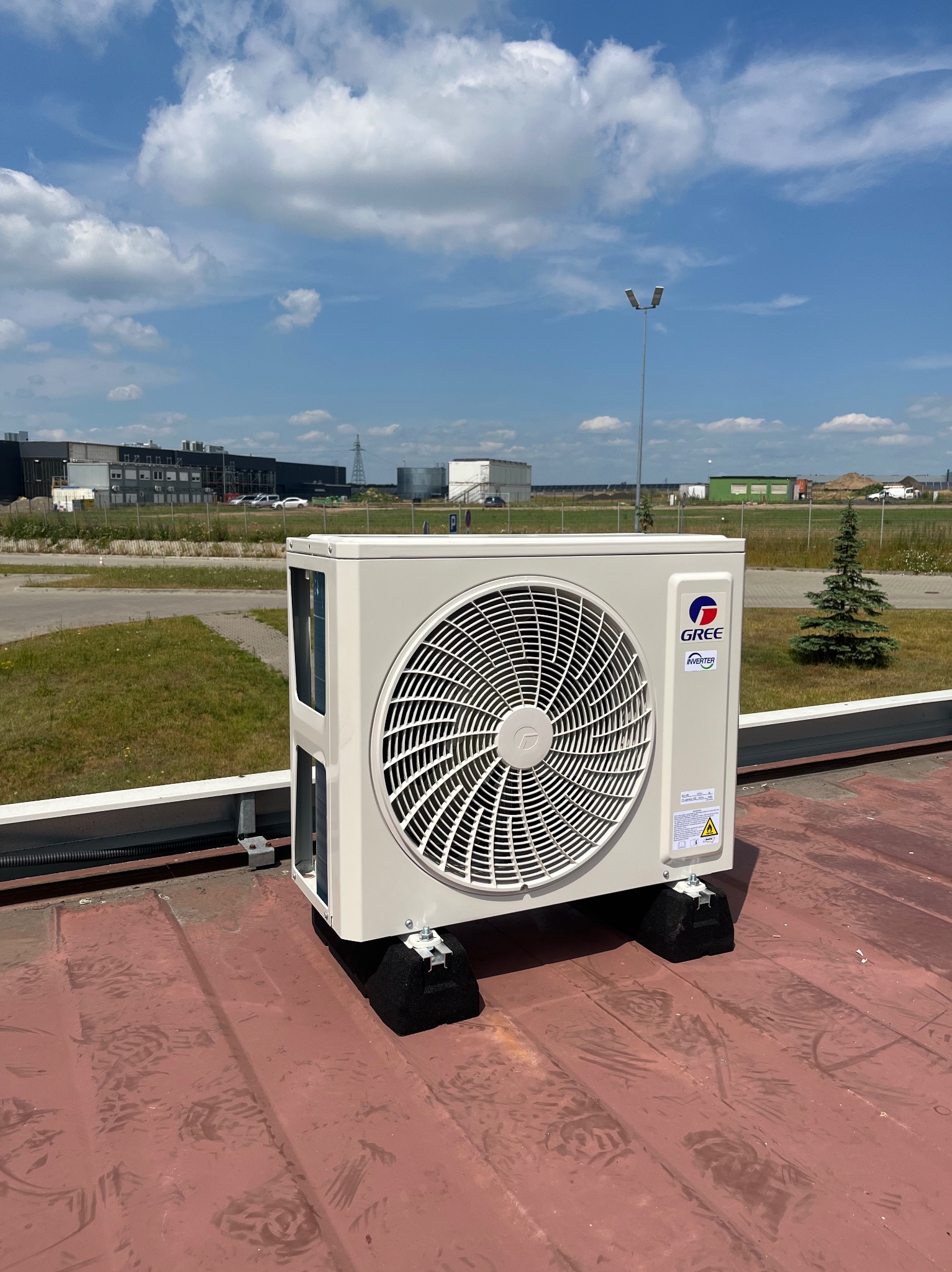 Zewnętrzna jednostka klimatyzacji marki Gree, zamontowana na dachu przemysłowego budynku, widoczna z bliska, z tłem błękitnego nieba i zabudowań przemysłowych.