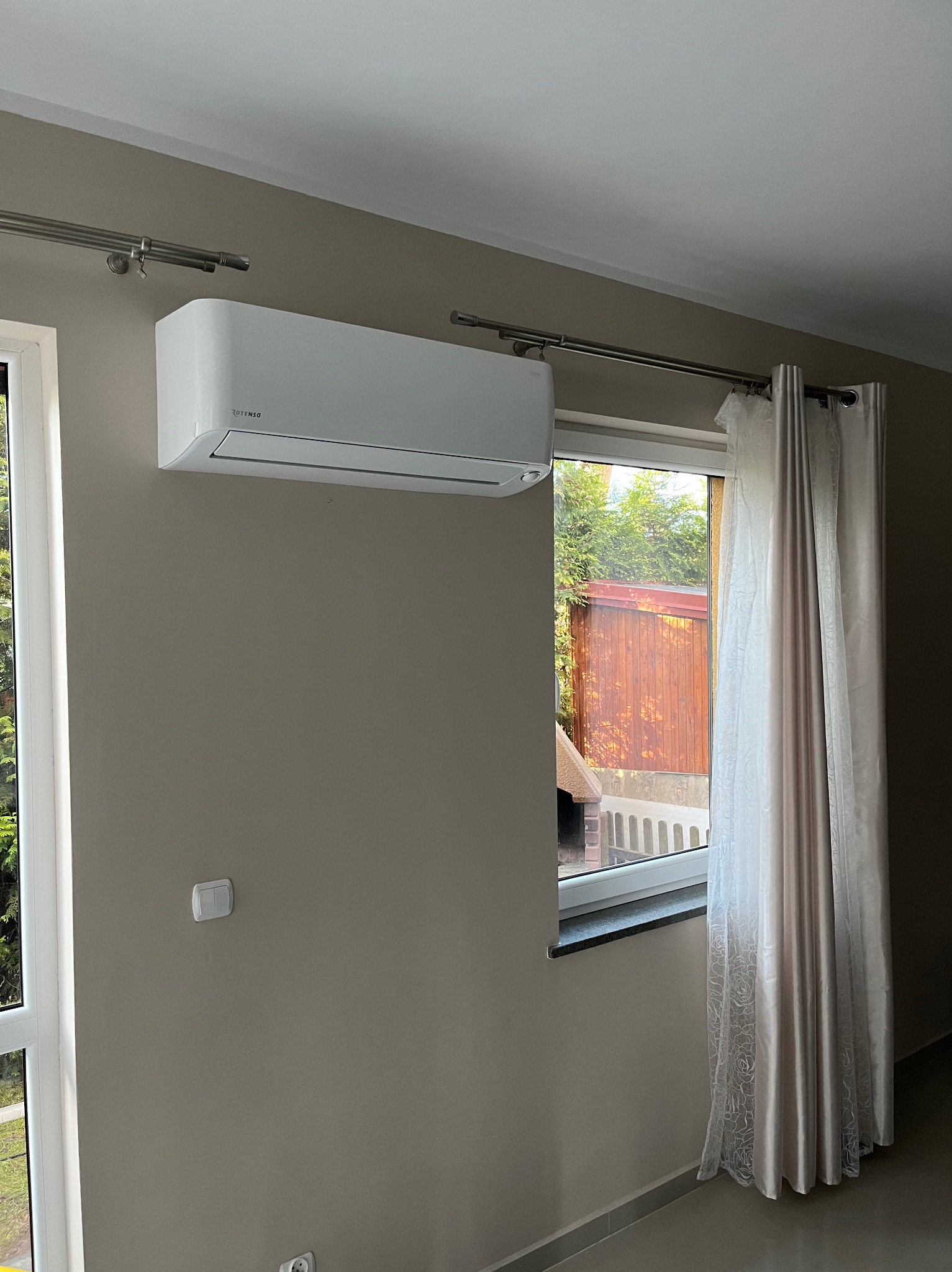 Biały klimatyzator Rotenso zamontowany na ścianie w pokoju z beżowymi ścianami, widok na ogród przez okno z zasłonami.
