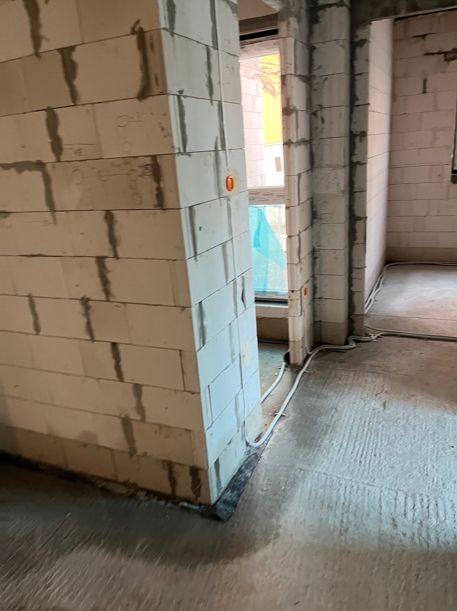 Surowy stan wnętrza budynku w trakcie budowy, widoczne ściany z betonu komórkowego, okno z niebieską folią ochronną i wystające przewody elektryczne.