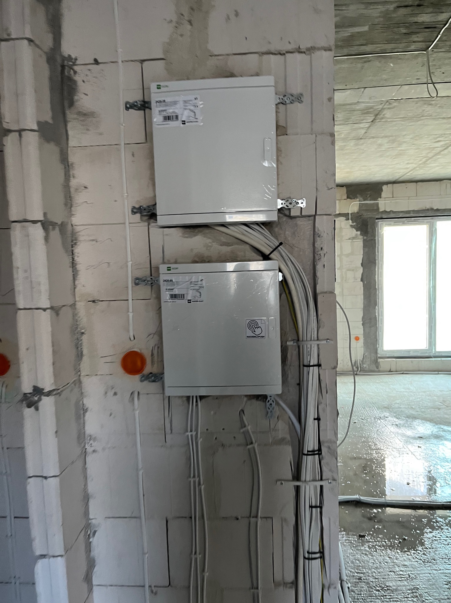 Dwa białe panele elektryczne zamontowane na surowej ścianie z betonu komórkowego, widoczne okablowanie, surowy stan wykończenia wnętrza.