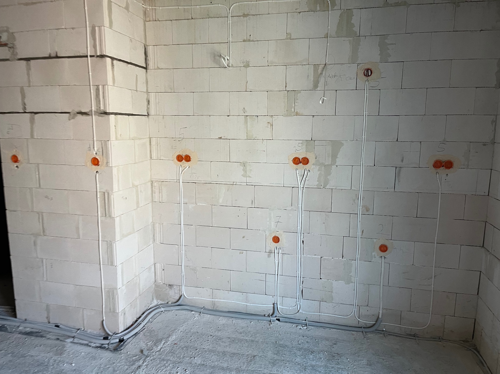 Szara ściana z betonu komórkowego w trakcie instalacji elektrycznej: widoczne kable w peszlach i puszki pod gniazdka, oznaczenia cyfrowe na ścianie.