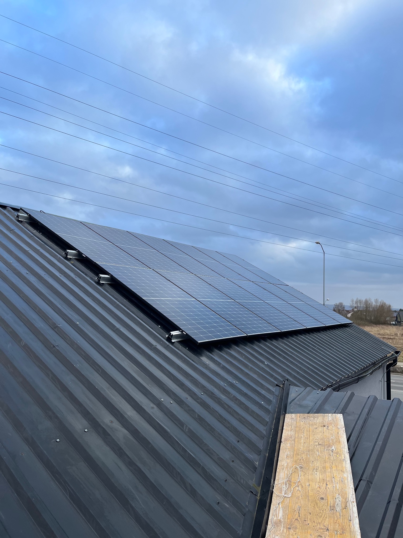 Panele fotowoltaiczne zamontowane na czarnym, blaszanym dachu domu jednorodzinnego, widok z góry, pochmurne niebo w tle.