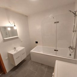 Usługi remontowo-budowlane - Brajan Cichecki - Biała łazienka po remoncie z wanną z parawanem, umywalką z szafką i szarą podłogą z płytek, widok z dołu.