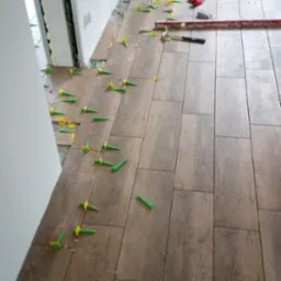 Układanie brązowych płytek podłogowych z użyciem zielono-żółtych klipsów poziomujących, widoczna poziomica i nóż do płytek.