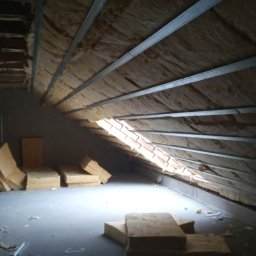 Usługi remontowo-budowlane - Brajan Cichecki - Poddasze w trakcie ocieplania, widoczne metalowe profile, wełna mineralna i fragment okna dachowego z ceglaną obudową, oświetlone naturalnym światłem.