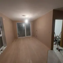 Pusta przestrzeń po remoncie z jasną, drewnianą podłogą, ścianami w odcieniach beżu i oknem zabezpieczonym folią, widok na otwarte drzwi z narzędziami i akcesoriami do sprzątania.