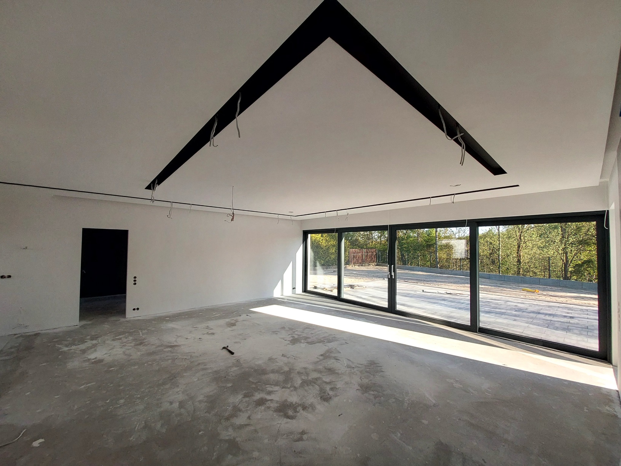 Wnętrze w stanie deweloperskim z surowym betonem na podłodze, dużym oknem przesuwnym, czarną, trójkątną konstrukcją dekoracyjną na suficie oraz wystającymi kablami.
