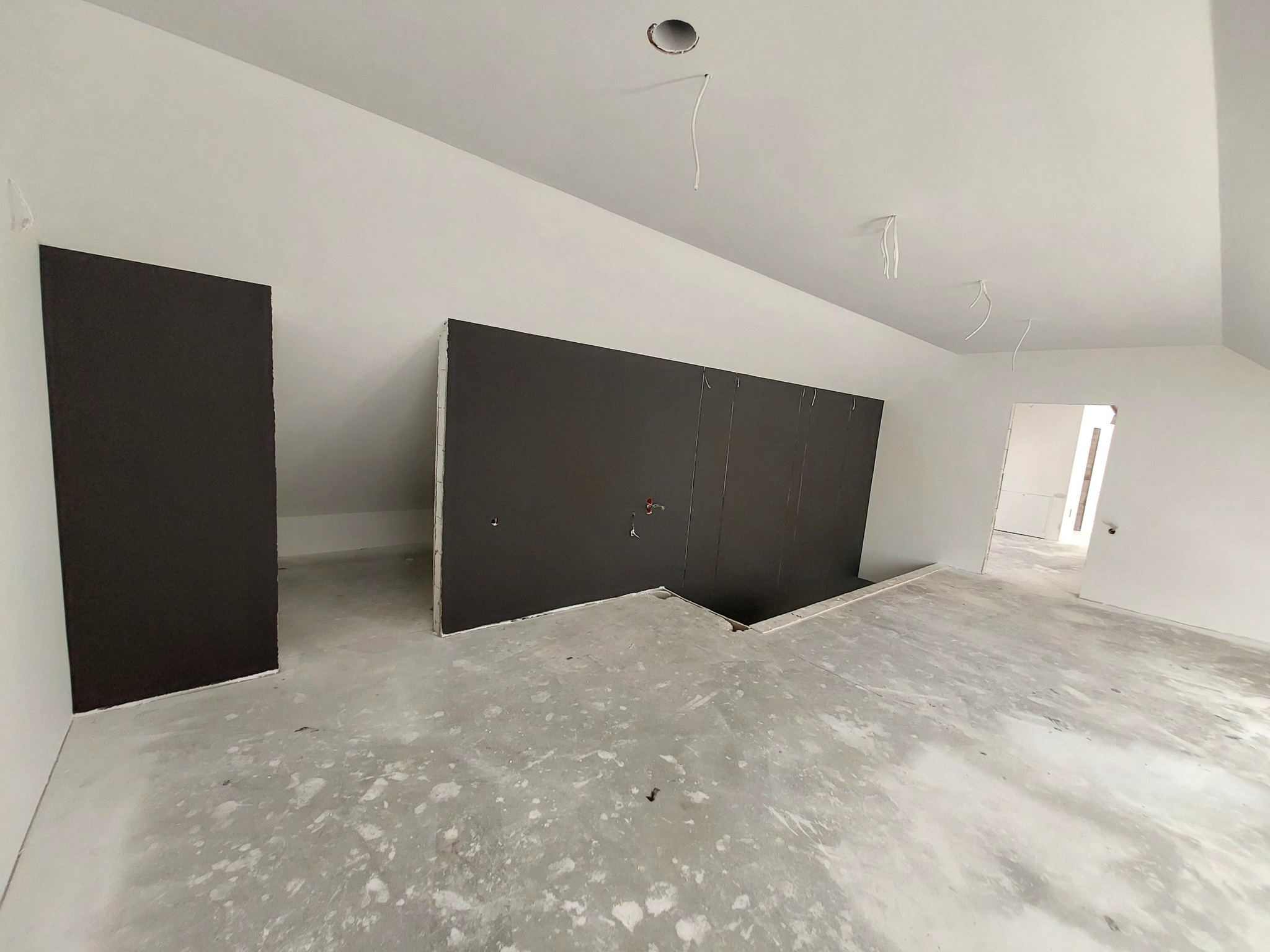 Poddasze w trakcie prac wykończeniowych, widoczne płyty kartonowo-gipsowe na ścianach, surowa podłoga i wystające kable z sufitu.