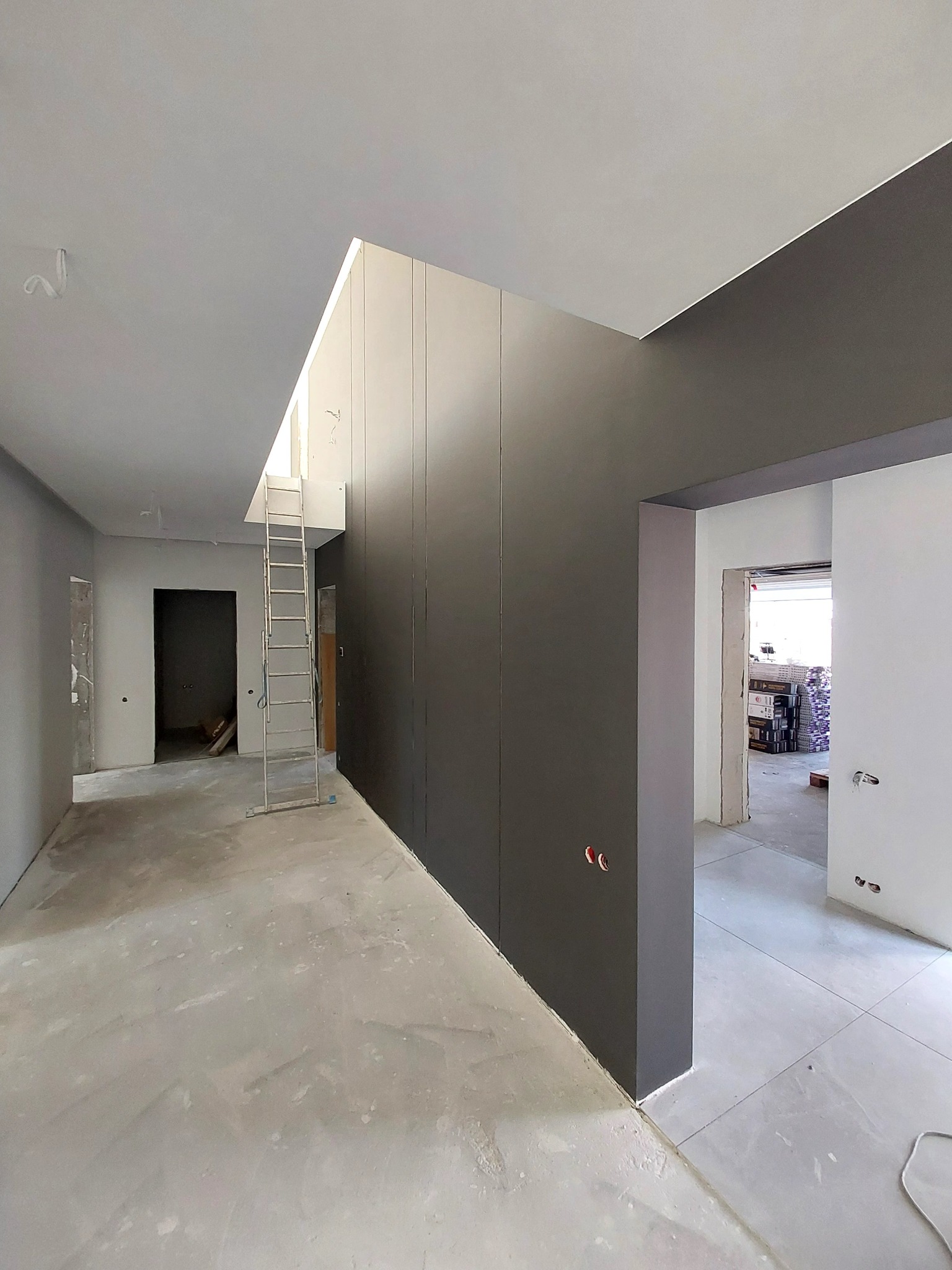 Wnętrze w trakcie prac wykończeniowych: szare ściany, surowy beton na podłodze i drabina oparta o ciemną zabudowę wnękową pod sufitem.