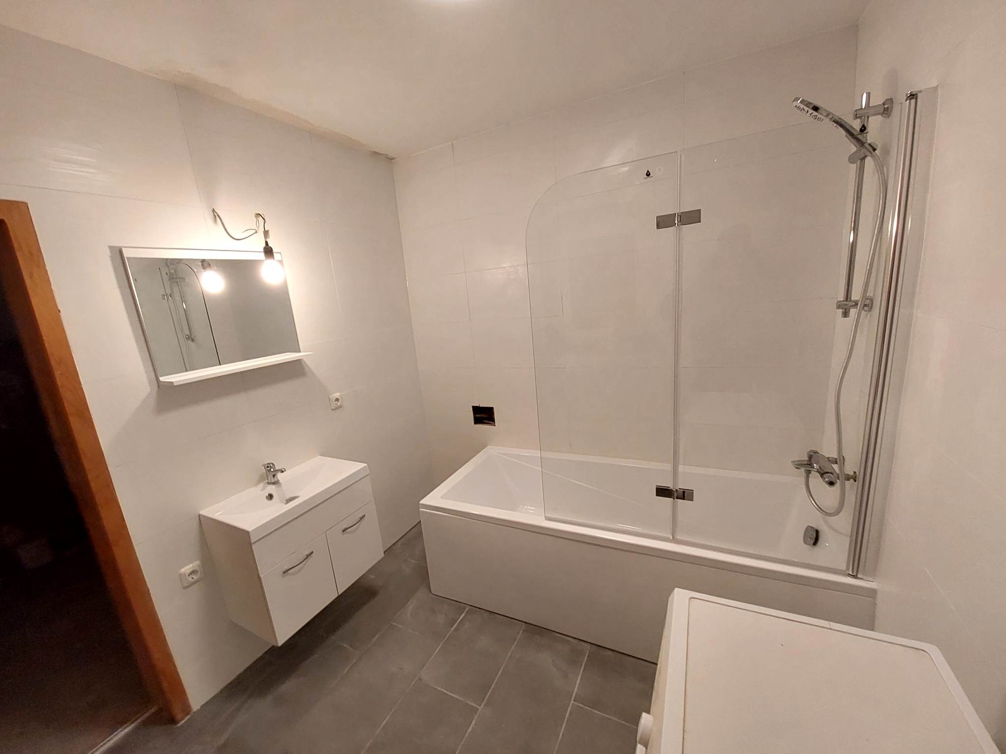 Biała łazienka po remoncie z wanną z parawanem, umywalką z szafką i szarą podłogą z płytek, widok z dołu.