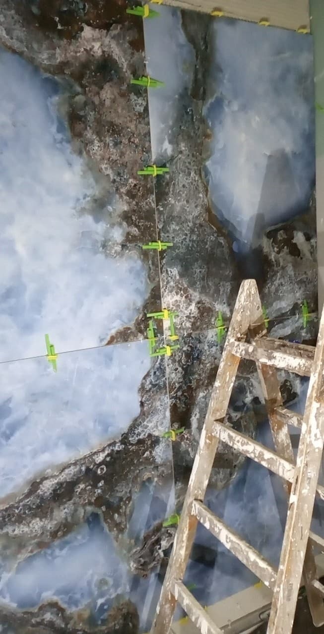 Montaż płytek imitujących marmur na ścianie, widoczne zielone krzyżyki dystansowe i drabina malarska.