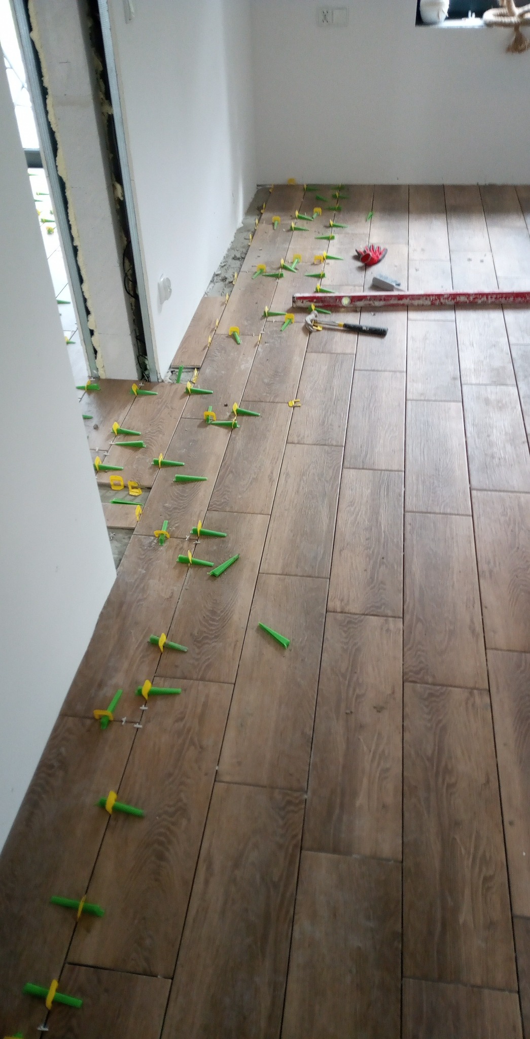 Układanie brązowych płytek podłogowych z użyciem zielono-żółtych klipsów poziomujących, widoczna poziomica i nóż do płytek.