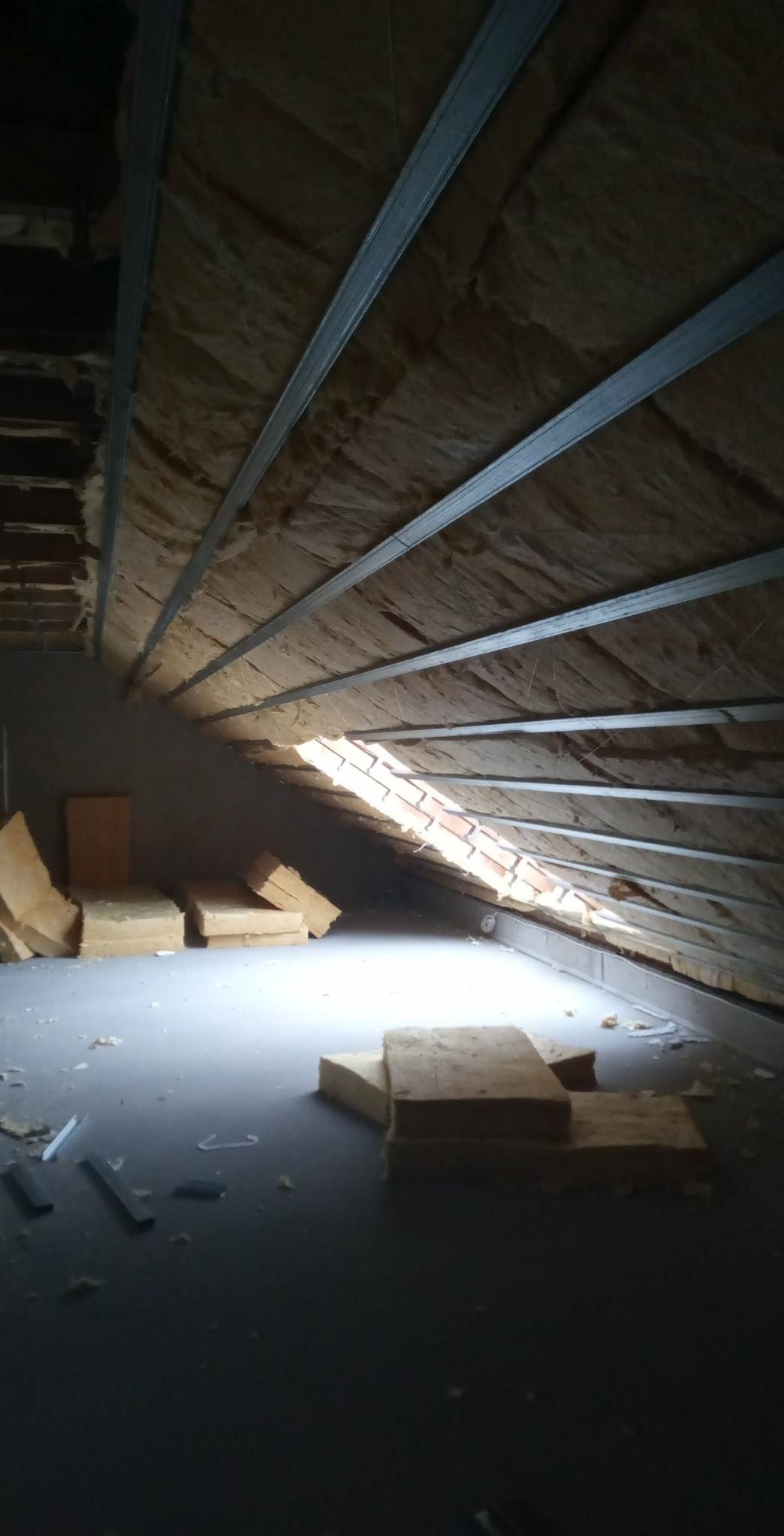 Poddasze w trakcie ocieplania, widoczne metalowe profile, wełna mineralna i fragment okna dachowego z ceglaną obudową, oświetlone naturalnym światłem.