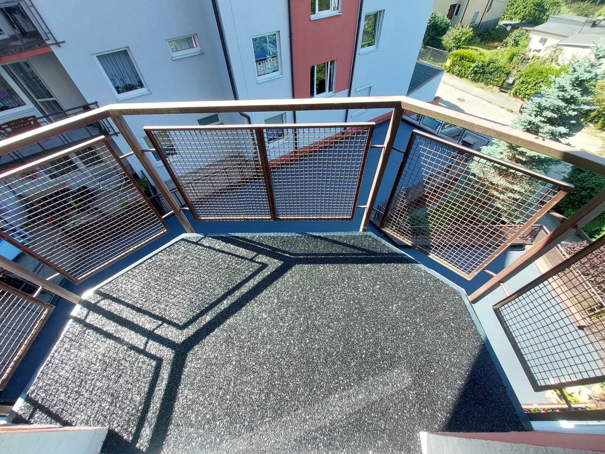 Widok z góry na zabudowany balkon z szarą, teksturowaną podłogą i metalową siatką ochronną, z widocznymi budynkami i zielenią w tle.
