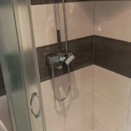 Kabina prysznicowa z chromowaną armaturą na tle płytek w odcieniach beżu i brązu, widoczna także pralka Inex.