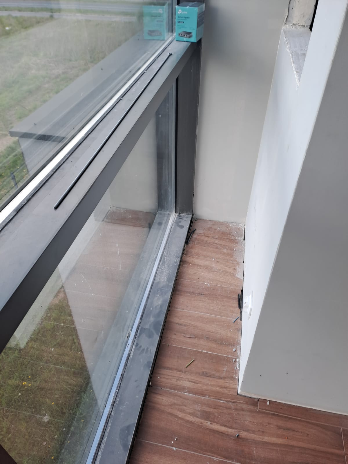 Kąt pomieszczenia z oknem, brudną ramą, fragmentem ściany i podłogą z płytek imitujących drewno. Na parapecie leżą dwa pudełka z urządzeniami elektronicznymi.