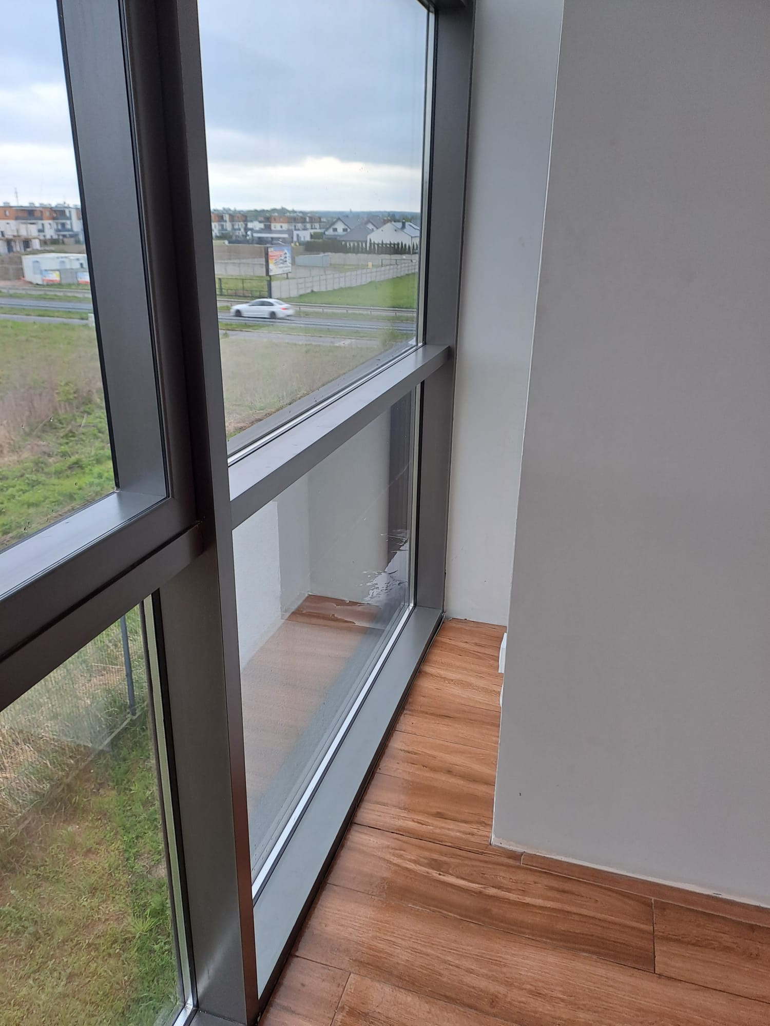 Brudne okno z widokiem na krajobraz miejski i zieleń, z wyraźnymi smugami i zaciekami na szybie, drewnopodobna podłoga.