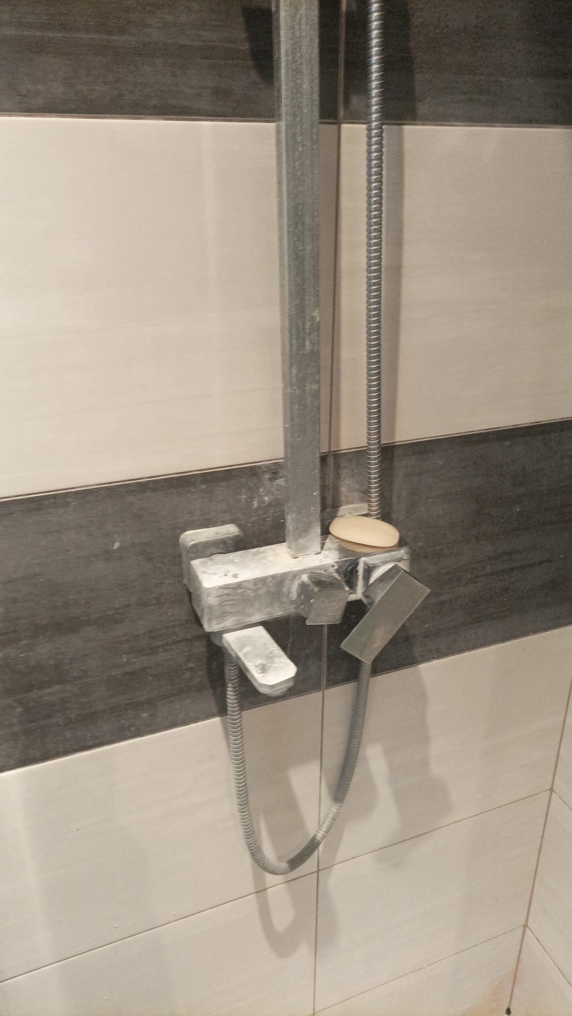 Zakamieniona bateria prysznicowa z mydłem na wierzchu, na tle białych i szarych płytek.