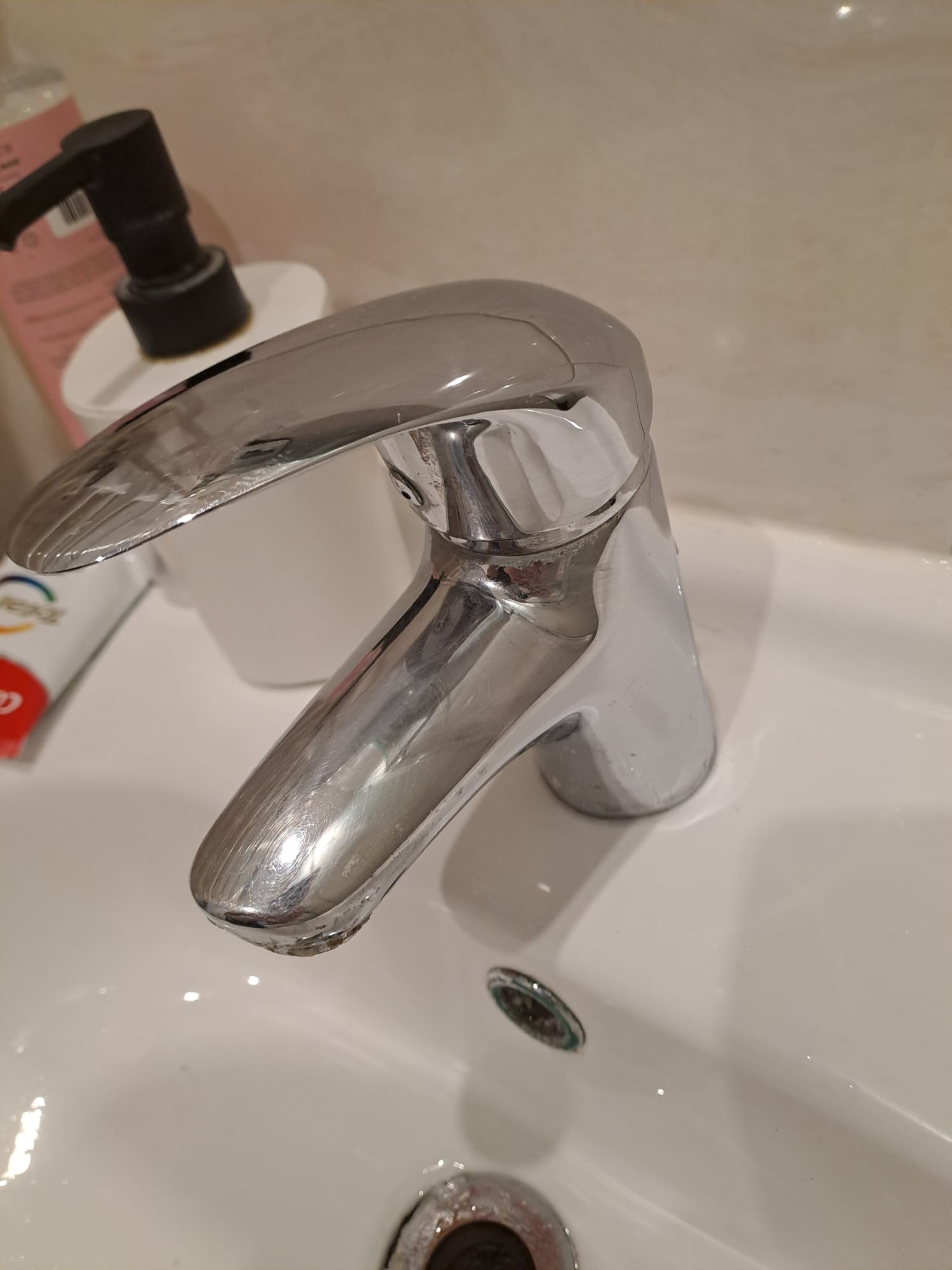 Błyszcząca chromowana bateria umywalkowa z widocznymi śladami osadu kamiennego, obok dozownik mydła i pasta do zębów, na białej umywalce.